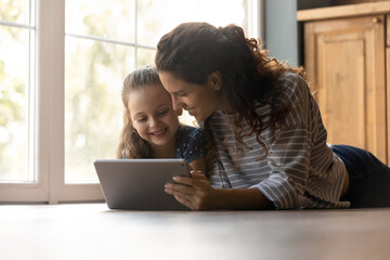 Caring young mom and little daughter lying on warm home floor have fun studying learning using modern pad gadget. Happy mother and small girl child look at tablet screen watching video online. © fizkes
