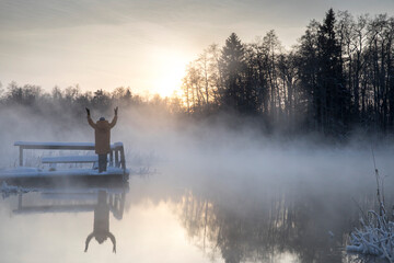 Obraz na płótnie Canvas Woman is standing on the wooden bridge and admire the sunset colored thick steam evaporating from the natural spring during the freezing cold winter day