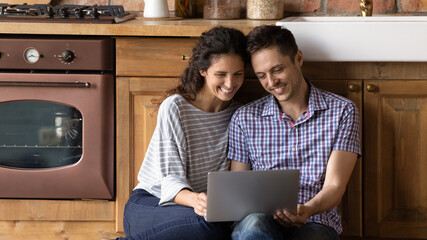 Wide banner view of happy young Caucasian man and woman relax in home kitchen browsing modern laptop together. Smiling millennial couple look at computer screen shopping online or watching video.