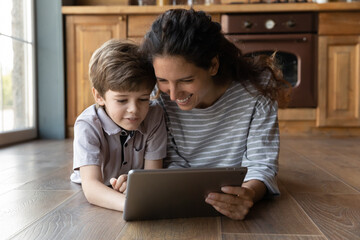 Smiling loving young Caucasian mom lying on warm floor using browsing tablet gadget with small son. Happy mother relax at home look at pad screen play online game or watch video with little boy child.