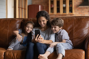 Happy young Caucasian mom sit relax on sofa with little children have fun using modern smartphone gadget. Smiling mother and small kids talk on video call or have webcam online communication on cell.