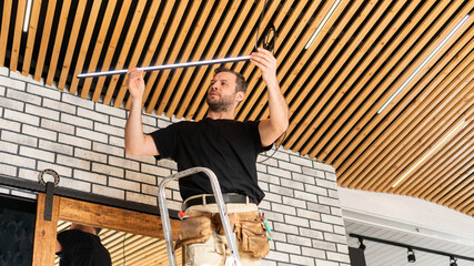 A male electrician changes lamps and repairs electrical wiring. A builder is installing a loft-style wooden plank ceiling. Installation of low-voltage LED lamps in the designer ceiling.