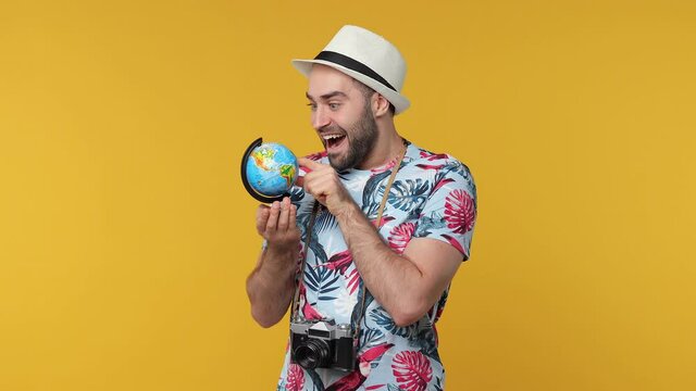 Cheerful traveler tourist young man in summer clothes hat isolated on yellow background. Passenger traveling on weekends. Air flight journey concept. Point finger on Earth world globe showing thumb up