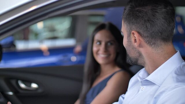 Close-up of cheerful woman and man enjoying sitting in dream car chosen for purchase in showroom. Satisfied male buyer happily holding steering wheel and stroking car while his wife sitting next