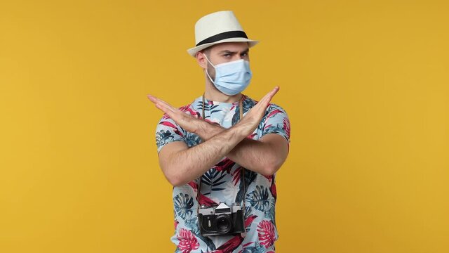 Funny traveler man point on face mask safe from coronavirus isolated on yellow background. Passenger travel on weekend. Air flight journey concept. Showing thumbs up hold crossed hands in stop gesture
