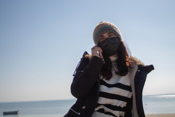 Beautiful girl with mask and hat next to the beach in winter talking on phone
