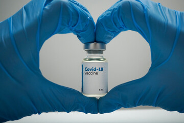 Heart-shaped hands in blue medical gloves holding a dose of coronavirus vaccine in a glass bottle. Liquid Covid-19 disease treatment for injections. Immunization against corona illness concept.
