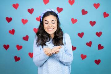 Young beautiful woman over blue background with red hearts Smiling with hands palms together receiving or giving gesture. Hold and protection