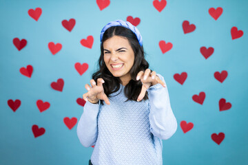 Young beautiful woman over blue background with red hearts smiling funny doing claw gesture as cat, aggressive and sexy expression