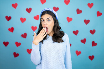 Young beautiful woman over blue background with red hearts covering mouth with hand, shocked and afraid for mistake. surprised expression
