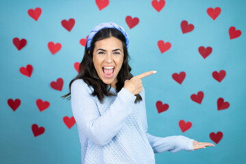 Young beautiful woman over blue background with red hearts surprised, showing and pointing something that is on her hand