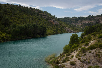 Fototapeta na wymiar Quiet blue water river in the Alarcon sickles full of green trees on a cloudy day, Alarcon, Cuenca, Spain