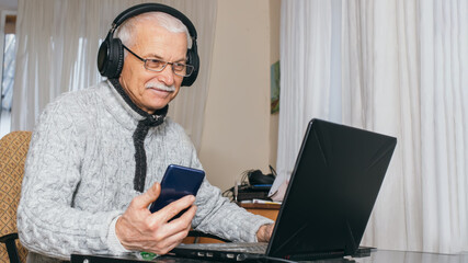 Handsome old man dressed in sweater and eyeglasses is using laptop and smiling while sitting at his work table at home. Grandpa chats in grandchildren on social media. senior man using phone