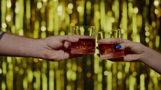 Two hands with glasses of cognac whiskey with ice cubes making cheers on gold background. Celebration of business success, Christmas, anniversary, birthday. Raising toast with glasses of brandy or rum