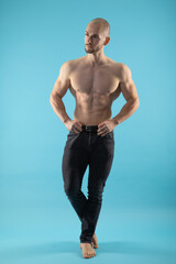 Fototapeta premium Muscular athletic guy dressed in black pants standing in blue Studio isolated looking to side and presenting his ripped six pack body shirtless