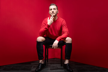 self addicted man in red studio sit in chair look straight into camera with his head high and fingers on chin