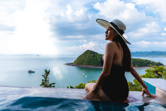 Asian women in black swimsuit and hat siting on side of infinity pool relaxing at sunset time with sea view during vacation retreat. Women happy and enjoy with holiday summer travel time. - Powered by Adobe