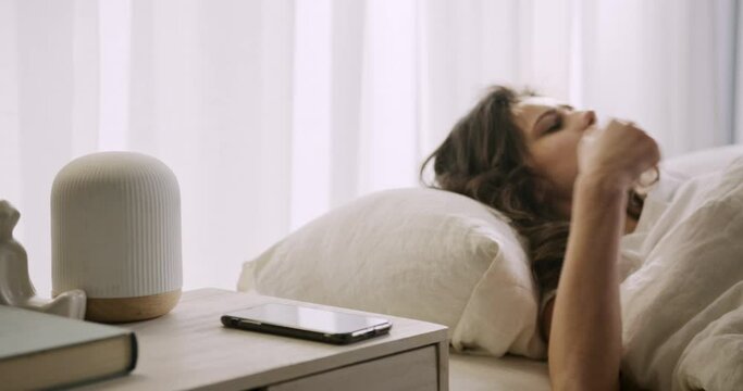 Young woman lying sound asleep in bed, wakes up suddenly to loud noise, looks irritated, switches off phone alarm clock, rolls over.