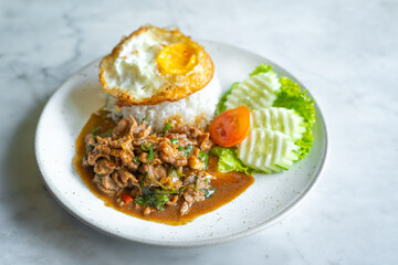Stir fried beef with basil and spicy chilly and rice topped with egg on the white dish on marble table. The delicious popular and famous favorite Thai food and street food.