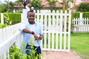 Healthy African American man holding a cup of coffee while leaning on the white picket fence in relaxation around the neighborhood with good green environment surrounding with copy space