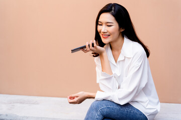 Young smiling Asian girl having conversation on smartphone while sitting on concrete bench with a desert sand colored wall in background wearing casual white shirt and jeans clothing