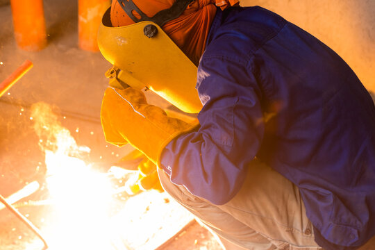 A Welder In Full Protective Gear Crouches Down And Welds Some Makeshift Tools. Hot Sparks Fly From The Welding Point.