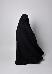 Full length portrait of pretty black haired woman wearing long dark gown nada cloak.  Standing pose facing away from the camera, against a  studio background.