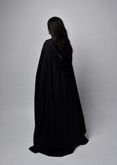 Full length portrait of pretty black haired woman wearing long dark gown nada cloak.  Standing pose facing away from the camera, against a  studio background.