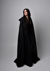 Naklejka premium Full length portrait of pretty black haired woman wearing long dark gown and a cloak. Standing pose facing away from the camera, against a studio background.