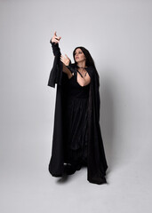 Full length portrait of pretty black haired woman wearing long dark gown and a cloak.  Standing pose facing away from the camera, against a  studio background.