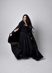 Naklejka premium Full length portrait of pretty black haired woman wearing long dark gown and a cloak. Standing pose facing away from the camera, against a studio background.