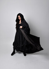 Full length portrait of pretty black haired woman wearing long dark gown and a cloak.  Standing pose facing away from the camera, against a  studio background.