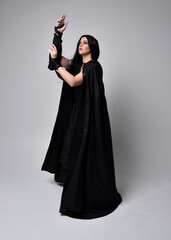 Full length portrait of pretty black haired woman wearing long dark gown and a cloak.  Standing pose facing away from the camera, against a  studio background.