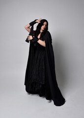 Naklejka premium Full length portrait of pretty black haired woman wearing long dark gown and a cloak. Standing pose facing away from the camera, against a studio background.