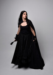 Naklejka premium Full length portrait of pretty black haired woman wearing long dark gown and a cloak. Standing pose facing away from the camera, against a studio background.