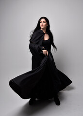 Naklejka premium Full length portrait of pretty black haired woman wearing long dark gown and a cloak. Standing pose facing away from the camera, against a studio background.