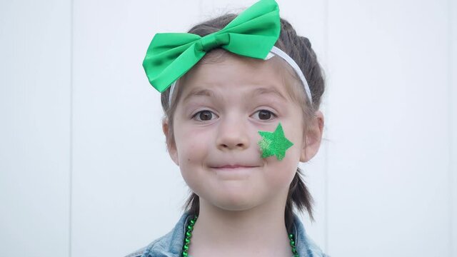 Cute happy brown eyes child with green bow on head looking at the camera, hiding green star on her cheek and smiling, celebrating saint patrick's day, white wall background. Close up
