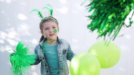 Girl with green funny horns on head and star on cheek holds two green balloons and pom poms, jumps and smiles looking at the camera, celebrating saint patrick's day. White wall background