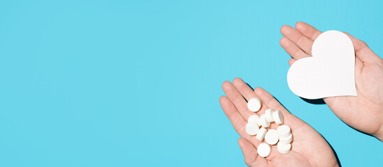 Hands holding a bunch of pills and a paper heart on a blue background, top view. Medical banner, copy space. Heart disease treatment, health care concept