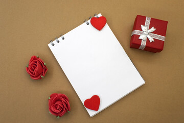 Among two decorative flowers in the form of roses and a red gift box is a notebook with a clean white sheet. Festive background. Template for the congratulations