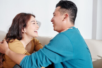 Happy young Asian boyfriend and girlfriend sitting on sofa, hugging and looking at each other