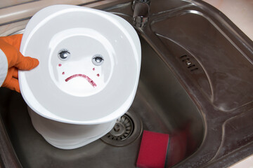 Poorly washed dishes in kitchen sink. Female hand holding white plate with eyes and sad emotion.