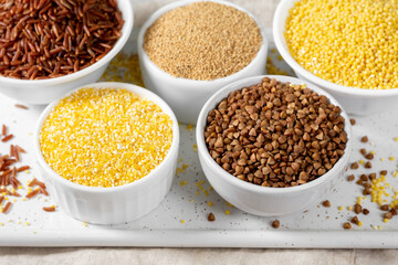 White and red rice, buckwheat, amaranth seeds, corn groats, quinoa and millet in white bowls on the light gray kitchen table. Gluten-free cereals