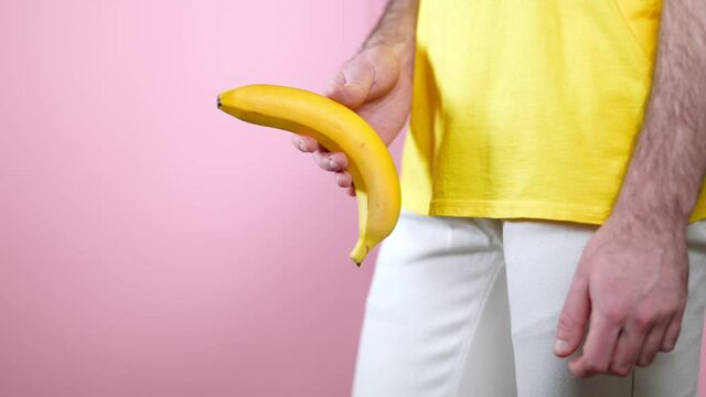 A man shows a banana in his hands at the level of the genitals. Side view. Pink background. The concept of men's health and potency.