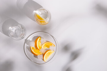 Drinking natural water and ice in beautiful glass glasses and slice of  fresh orange on plate on white background. Summer minimal flat lay with hard shadows.
