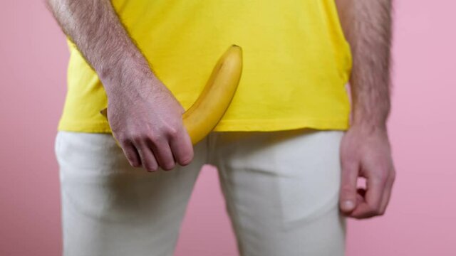 Man holds a lowered banana in his hand at the level of the genitals. A woman's hand runs a finger over a banana and it rises up. Pink background. The concept of men's health.