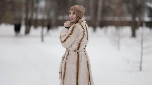 An attractive fashionable blonde in a beautiful fur coat and a beige hat poses against the background of a winter park. Model on a snowy background of winter weather.