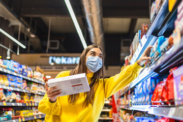 Fototapeta premium Social distancing in a supermarket. A young woman in a disposable face mask buying food and putting them in a grocery basket. Shopping during the Coronavirus Covid-19 epidemic 2020