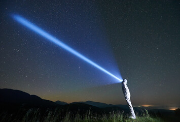 Cosmonaut illuminating fantastic night sky with flashlight. Cosmonaut wearing white space suit and helmet while standing in grassy valley. Concept of light, galaxy and astronautics.
