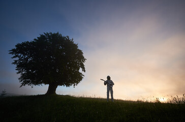 Horizontal snapshot of two silhouettes on purple sky background, of a spaceman in space suit playing guitar and a big old tree on the right. Concept of music, cosmonautics and nature.